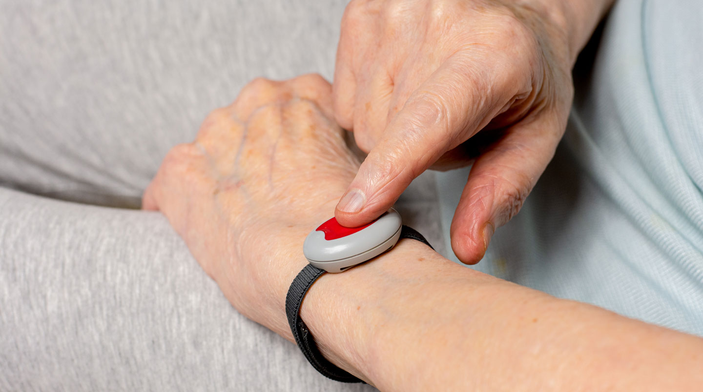 Homme senior en chemise bleue testant un bouton d'appel d'urgence rouge dans une salle de bain adaptée PMR avec douche à l'italienne, barres d'appui chromées et carrelage blanc
